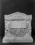 Cinerarium with Genii at the corners holding a garland, name plate with medallion portrait below it, and separate lid. 