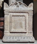 Roman cinerarium and lid: between double fillet and compressed waterleaf mouldings is a large name plate with similar borders flanked by two enriched Tuscan pilasters. The lid has a curved pediment ornamented with an eagle with wings half spread and an antefix on each of the four corners with enriched borders below on three sides. There are large palmettes carved on the ends / sides of the cinerarium. 
