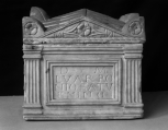 A Roman funerary urn (cinerarium) with fluted Tuscan pilasters at the corners and a central name plate framed with double mouldng strips.  At the corners of the lid are antefixae decorated with inset rosettes and there is a  similar rosette flanked by stylised foliage within the triangular pediment. 
